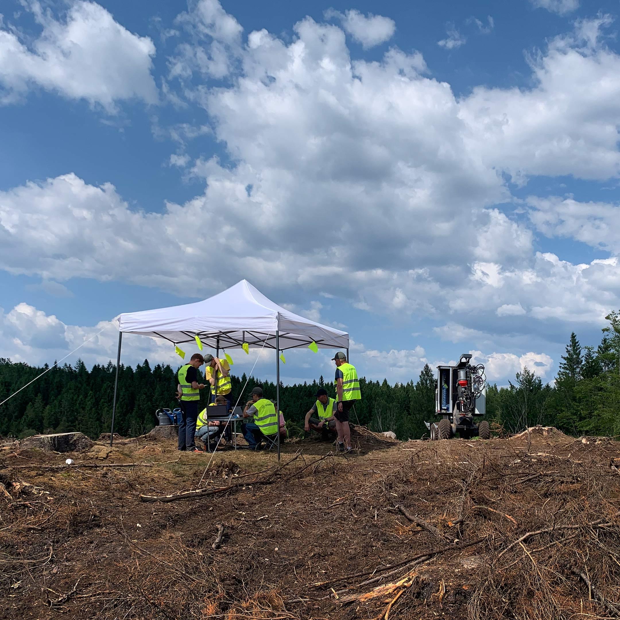 Fem personer som står under ett uppsatt soltält ute i skogen. Till höger om tältet står en planteringsrobot.
